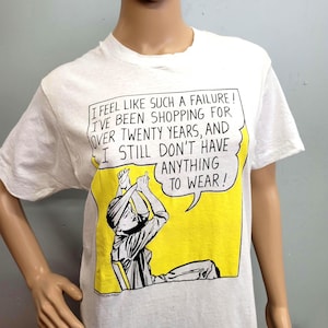 May include: White t-shirt with a comic-style graphic in yellow and black. The graphic features a person with their hands raised and a speech bubble that reads, "I FEEL LIKE SUCH A FAILURE! I'VE BEEN SHOPPING FOR OVER TWENTY YEARS, AND I STILL DON'T HAVE ANYTHING TO WEAR!"