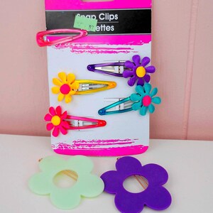 May include: A package of snap clips with the text "basic solutions" and "Snap Clips". The package contains four snap clips with flower accents in various colors: pink, yellow, purple, and teal. Two additional flower-shaped hair accessories are also visible.