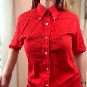 May include: A bright red short-sleeved button-down shirt featuring a pointed collar with small buttons at the tips, a central vertical row of white buttons, and a single patch pocket on the left side of the chest. The fabric has a visible texture.