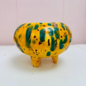 Puede incluir: Pequeña maceta redonda de cerámica con tres patas cortas. La maceta es principalmente amarilla con manchas irregulares de color verde azulado y negro. El interior no es visible. Adecuada para plantas pequeñas o suculentas.