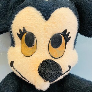 May include: Close-up of a vintage plush toy featuring a mouse character. The toy has a cream-colored face with large, round eyes, black eyelashes, and a black nose. The ears and the rest of the body are black.
