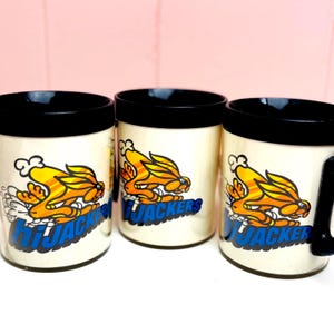 Könnte beinhalten: Drei cremefarbene Keramikbecher mit schwarzen Rändern und Henkeln. Jeder Becher zeigt eine Cartoon-Grafik einer gelb-orangenen Kreatur mit dem Wort "HIJACKERS" in Blau. Die Becher werden vor einem rosa Hintergrund präsentiert.