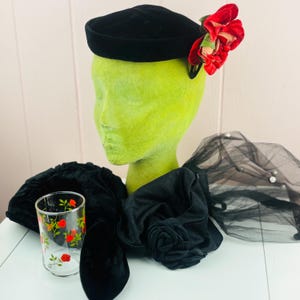 May include: A black velvet pillbox hat adorned with red fabric flowers, a black fabric headpiece featuring a central fabric rose, a black tulle veil with small white beads, and a clear glass tumbler decorated with a pattern of small red roses.
