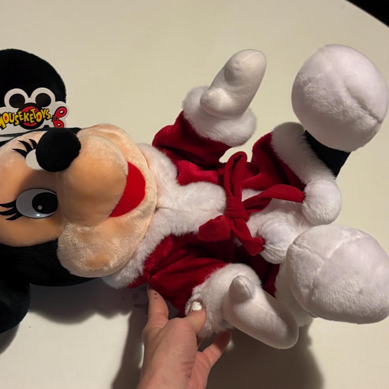Pode incluir: Um brinquedo de pel&uacute;cia da Minnie Mouse vestida com uma roupa de Papai Noel. O brinquedo &eacute; principalmente vermelho e branco, com detalhes em preto e um la&ccedil;o. O brinquedo tem uma etiqueta que diz "Mouseketoys".