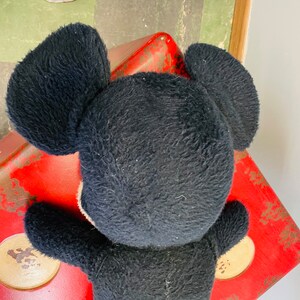 May include: A vintage black plush toy with large, round ears and a white face. The toy is positioned on a distressed red surface, suggesting it is an older item. The plush material appears worn, adding to its antique charm. The toy's design is reminiscent of a classic cartoon character.