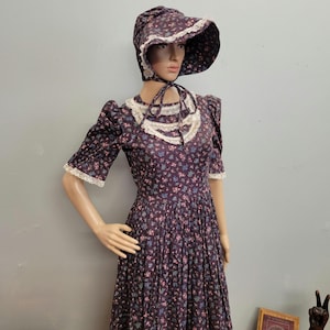 May include: A dark purple floral print dress and matching bonnet. The dress features short sleeves with white lace trim, a high neckline with lace detailing, and a full skirt. The bonnet has a wide brim and ties under the chin.