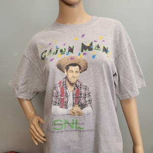Puede incluir: Camiseta gris con un gráfico de un hombre con sombrero de paja y tirantes. El texto dice "Cajun Man" y "SNL SATURDAYNIGHTLIVE". La camiseta tiene cuello redondo y mangas cortas.