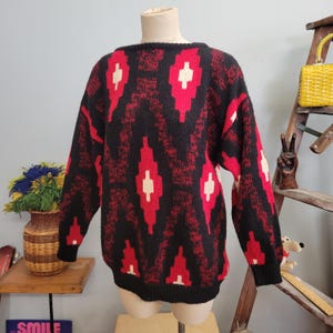 May include: A black sweater with a red and white geometric pattern. The long-sleeved sweater is displayed on a mannequin. The pattern includes red diamond shapes with white accents, set against a black background. The sweater has a crew neck and black cuffs.