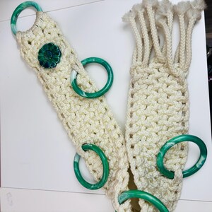 May include: A cream-colored macrame strap featuring a series of marbled green circular rings integrated into the woven cord pattern. A single dark green flower-shaped button is attached to the top section, and the end features loose cord fringe.
