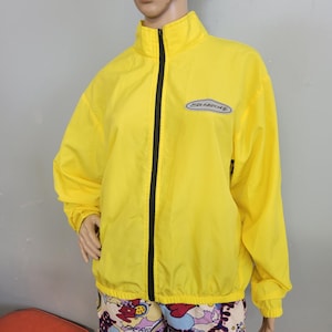 May include: Bright yellow zip-up jacket with a black zipper and a small logo on the chest. The jacket has a high collar and long sleeves. The jacket is made of a lightweight material.