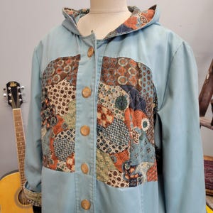 May include: A light blue hooded jacket featuring a front panel with a multi-colored quilted patchwork pattern in shades of orange, brown, green, and blue. The jacket has a button-down front with five round brown buttons and a matching patterned hood lining.