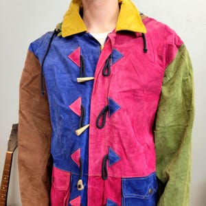Op de afbeelding: Een kleurrijke patchworkjas met een gele kraag en capuchon. De jas heeft panelen van bruin, blauw, roze en groen suède. Het heeft knevelsluitingen en een rits. Een uniek en opvallend kledingstuk.