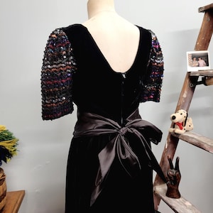 May include: A black velvet dress with a satin bow at the waist. The sleeves are adorned with colorful sequins. The dress has a V-neckline and a zipper down the back. The dress is displayed on a mannequin.