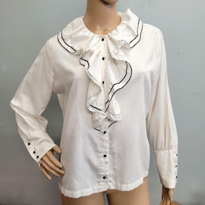 Peut inclure: Chemisier blanc avec col et devant à volants, soulignés de noir. La chemise à manches longues comporte des boutons noirs sur le devant et aux poignets. Vêtement d'inspiration vintage.