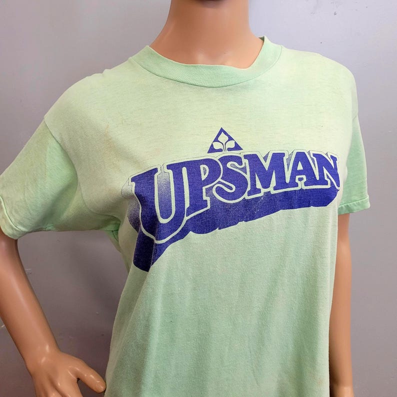 May include: A light green t-shirt with the word "UPSMAN" in blue, stylized text. Above the text is a blue triangle with a white design. The shirt has short sleeves and a crew neck.
