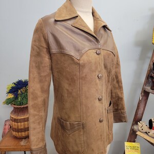 May include: A brown suede jacket with a brown leather yoke and pockets. The jacket has a button front closure and long sleeves.