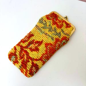 May include: A vintage eyeglass case with a woven design. The case features a yellow background with red and black floral patterns. It has a gold-colored metal clasp.