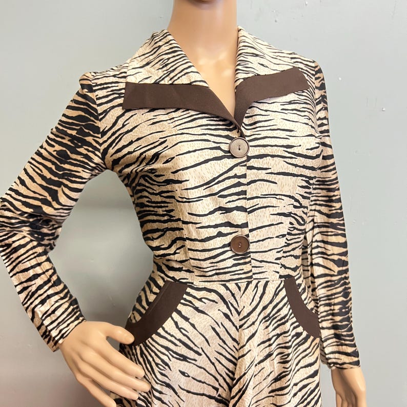 May include: A long-sleeved dress featuring a tan and black tiger stripe pattern. The garment includes a pointed collar with solid brown lapel accents, two brown buttons down the center front, and two brown pocket flaps at the waist area.