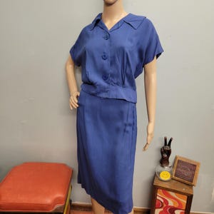 May include: A vintage blue two-piece outfit. It features a collared, button-down top with short sleeves and a matching knee-length skirt. The outfit is displayed on a mannequin, highlighting its classic design and tailored fit. The fabric has a slight sheen.