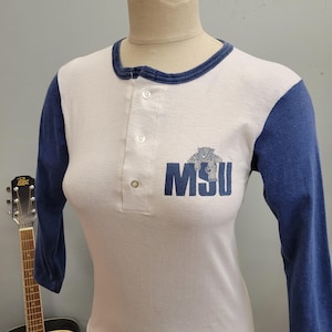 Könnte beinhalten: Weißes und blaues Raglan-Shirt mit langen Ärmeln. Das Shirt hat einen Rundhalsausschnitt und eine Knopfleiste mit drei Knöpfen. Auf der Vorderseite des Shirts sind die Buchstaben "MSU" mit einer Raubkatzengrafik abgebildet. Die Ärmel sind blau.
