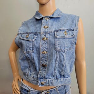 May include: A light blue denim vest featuring a classic collar, a front button-down closure with metallic buttons, and two chest pockets with buttoned flaps. The sleeveless garment has a cropped length and visible stitching details throughout.