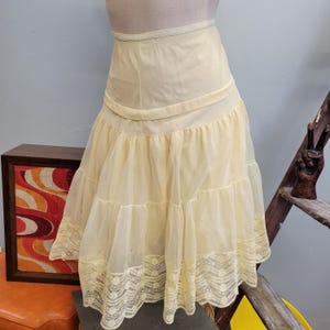 May include: A vintage, pale yellow petticoat with a tiered skirt and lace trim. The petticoat is made of a sheer fabric and has a fitted waist. The lace trim is a matching pale yellow color. The petticoat is displayed on a mannequin.