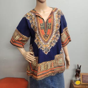 May include: A navy blue short-sleeved shirt featuring a prominent V-neckline and a vibrant, multi-colored geometric pattern in shades of orange, yellow, and cream. The intricate design covers the chest, sleeves, and hem, creating a bold visual contrast against the dark blue fabric base.