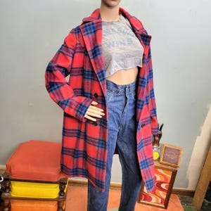 May include: A red and blue plaid coat with a double-breasted design. The coat is displayed on a mannequin, paired with a gray crop top and blue jeans. The coat has a classic, longline silhouette.