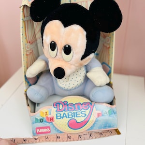 May include: A vintage Playskool Disney Babies Mickey Mouse plush toy in its original box. The plush Mickey Mouse is wearing a blue outfit and bib with white polka dots. The box features the Disney Babies logo and colorful graphics.