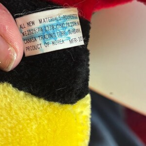 May include: Close-up of a plush toy with red, black, and yellow sections. A tag is visible with text: "ALL NEW MATERIALS-POLYESTER, CANASA TRADING CORP. BURBANK, PRODUCT OF KOREA".