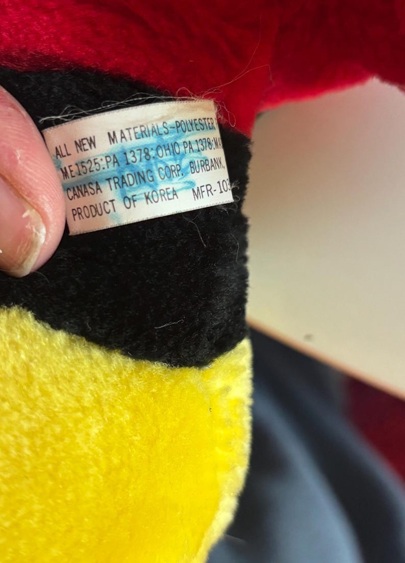 May include: Close-up of a plush toy with red, black, and yellow sections. A tag is visible with text: "ALL NEW MATERIALS-POLYESTER, CANASA TRADING CORP. BURBANK, PRODUCT OF KOREA".