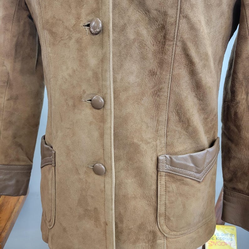 May include: A brown suede jacket with a brown leather trim and three buttons down the front. The jacket has a patch pocket on the left side and a flap pocket on the right side.