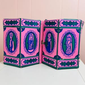 Könnte beinhalten: Eine vintage, pinkfarbene Faltbox mit grünen und blauen Akzenten. Die Box zeigt ovale Porträts von Frauen in blauen und grünen Rahmen. Dekorative Blumen- und Blättermuster zieren die Ränder.