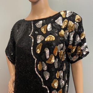 Puede incluir: Top negro con lentejuelas y mangas cortas. El top presenta un ribete de lentejuelas plateadas y un patrón de lentejuelas doradas, plateadas y negras en el hombro derecho. La prenda se exhibe en un maniquí.