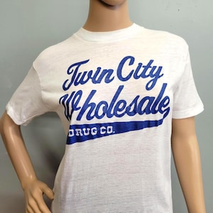 May include: A white short-sleeve t-shirt featuring blue screen-printed text that reads Twin City Wholesale DRUG CO. The graphic is arranged in a script font for the first two lines with a solid blue banner shape underneath the final line.