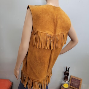 May include: A tan suede vest with fringe detailing along the bottom and across the upper back. The sleeveless vest is displayed on a mannequin, showcasing its vintage style and texture.