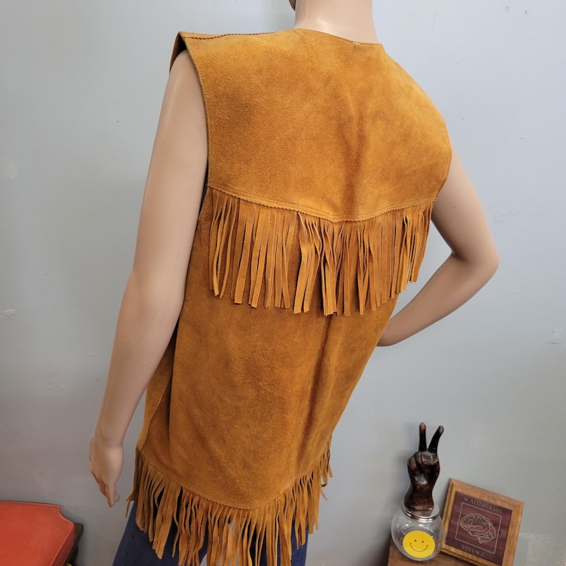 May include: A tan suede vest with fringe detailing along the bottom and across the upper back. The sleeveless vest is displayed on a mannequin, showcasing its vintage style and texture.