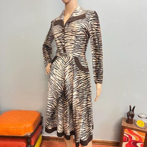 May include: A long-sleeved dress featuring a tan and black tiger stripe pattern. The dress has a collared neckline, two brown buttons down the front, and two brown pocket accents at the waist. The hem and collar are finished with solid brown trim.