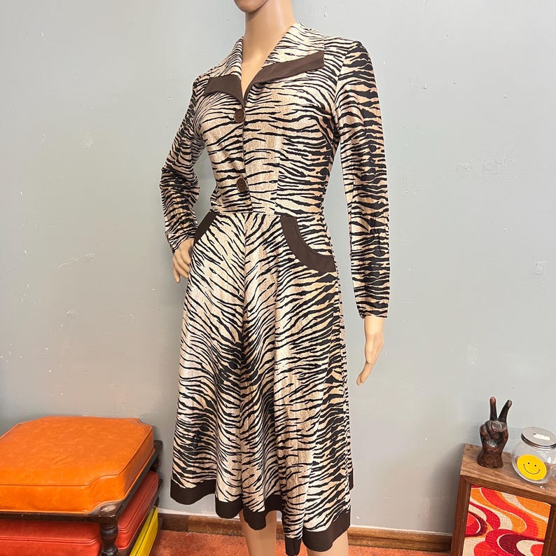 May include: A long-sleeved dress featuring a tan and black tiger stripe pattern. The dress has a collared neckline, two brown buttons down the front, and two brown pocket accents at the waist. The hem and collar are finished with solid brown trim.