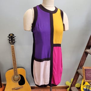 May include: A sleeveless, color-block dress in a Mondrian-inspired design. The dress features panels of purple, yellow, pink, and off-white, outlined in black. The dress is displayed on a mannequin.