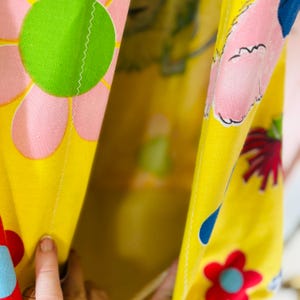 May include: Close-up of a yellow floral fabric. The design includes large pink and green flowers, with a white zigzag stitch along the seams. The fabric has a bright, cheerful appearance, with a vintage aesthetic.