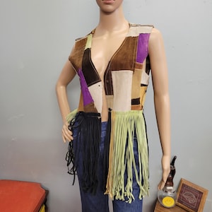 May include: A sleeveless vest constructed from various rectangular patches of suede in shades of brown, tan, and purple. The garment features a deep V-neckline and a long fringe hemline in contrasting black and light green strips of suede material.