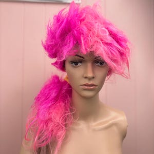 May include: A bright pink wig with a voluminous, textured style and a ponytail. The wig is displayed on a mannequin head, showcasing its vibrant color and playful design. The wig's texture appears fluffy and layered.