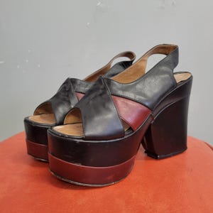 May include: A pair of vintage platform sandals. The sandals feature a black leather upper with a burgundy accent and a thick platform sole. The heels are blocky and the sandals have a slingback strap. The shoes are displayed on a red surface.