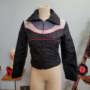 May include: Black, quilted jacket with a stand-up collar and a zipper closure. The jacket features a color block design with gray, red, and blue accents. The jacket is displayed on a mannequin.