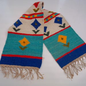 May include: A woven scarf featuring an off-white base with geometric patterns in turquoise, royal blue, red, yellow, and dark green. The design includes diamond shapes and floral motifs, finished with thin, light beige fringe at both ends.