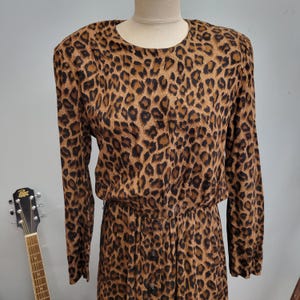 May include: A vintage leopard print dress with long sleeves and a button-down front. The dress features a brown and black animal print pattern and is displayed on a mannequin. The dress has a round neckline and shoulder pads.