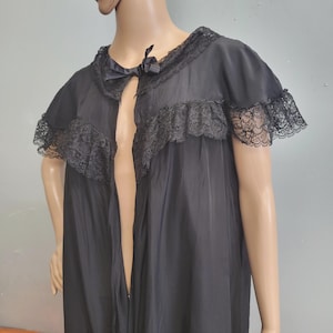 Puede incluir: Una bata negra transparente con mangas cortas adornadas con encaje con volantes, un cuello a juego con encaje y un pequeño lazo negro en el escote. La prenda está confeccionada con un tejido ligero y semitransparente de silueta fluida.