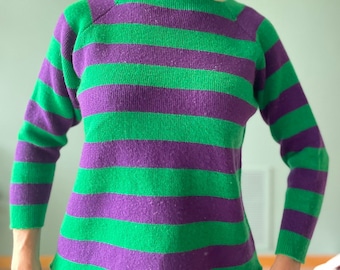 May include: A green and purple striped sweater with a boat neck and long sleeves. The sweater features horizontal stripes of green and purple. The sleeves are long and the neckline is a boat neck style. The sweater is made of a soft, knitted material.