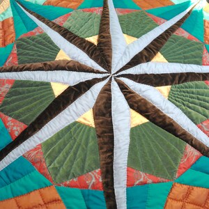 Op de afbeelding: Een gewatteerd textiel met een centraal kompasroosontwerp met afwisselende lichtblauwe en donkerbruine fluwelen punten. Het omliggende patroon bevat geometrische secties in groenblauw, oranje en olijfgroen met gedetailleerd stikwerk.